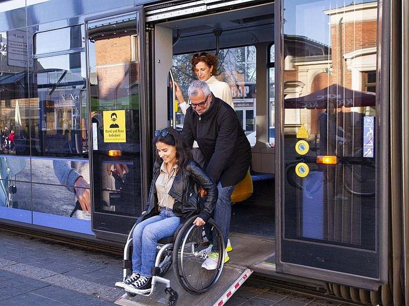 A man helps a woman in a wheelchair to leave a bus via the bus's own ramp.
