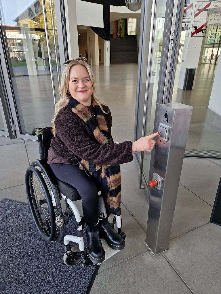 Frau im Rollstuhl zeigt auf Eingangstüröffnertaste