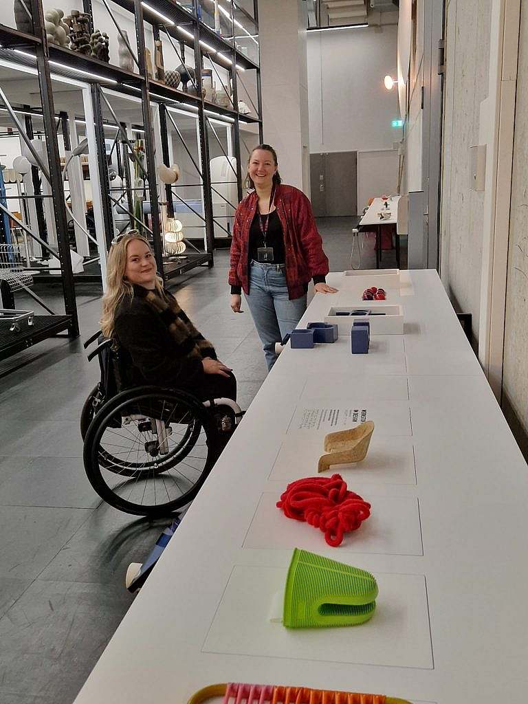 Frau im Rollstuhl mit Begleiterin vor einem Tisch mit Ausstellungsobjekten