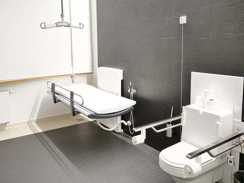 Innenraum von „Toilette für alle“, WC mit Griffen, höhenverstellbar, Pflegeliege ebenfalls höhenverstellbar und Deckenlift