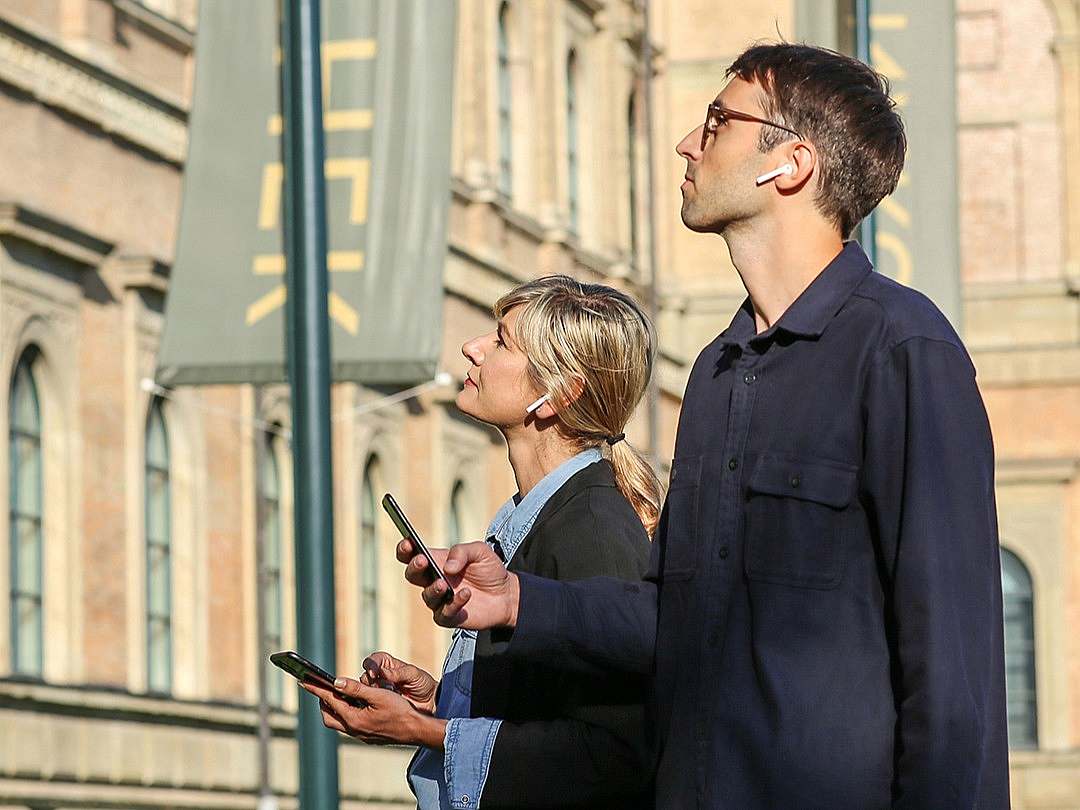 Two people wearing headphones and holding smartphones in front of a building