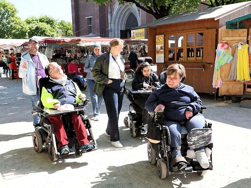 Besuchergruppe mit mehreren Fußgängern und Rollstuhlfahrern vor Dult-Ständen, im Hinterrgrund der Kirchenbau