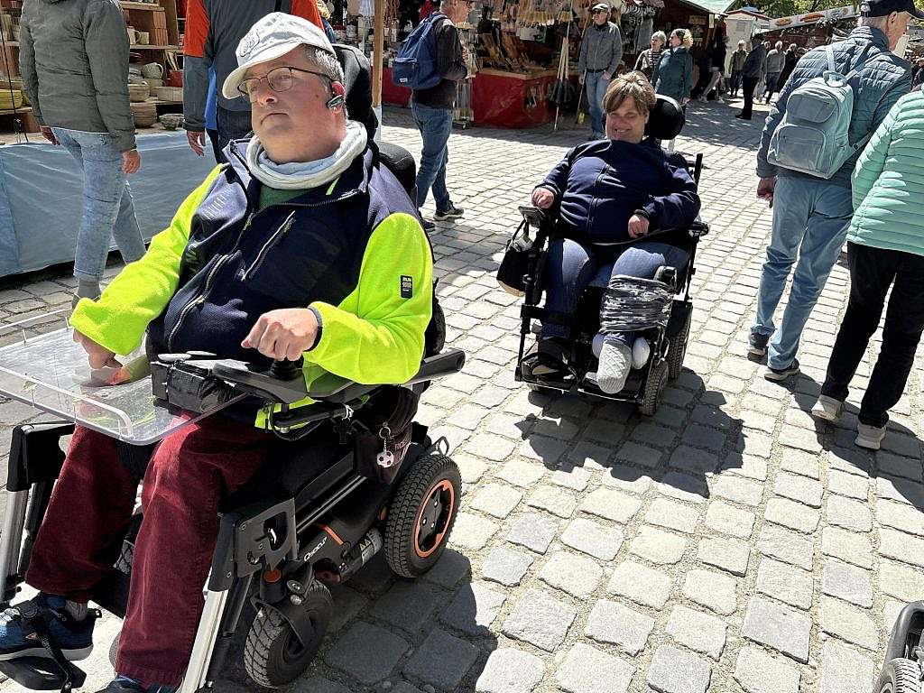 Two people in electric wheelchairs navigating independently between market stalls on cobblestones