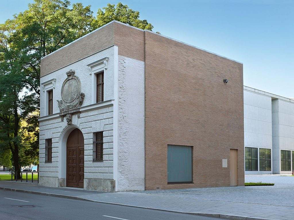 Former barracks, viewed from the front and the side. Front: a white-grey façade with four windows, a sort of panel at the top and an arched door at the bottom. Side: a brown façade with one window and a narrow door