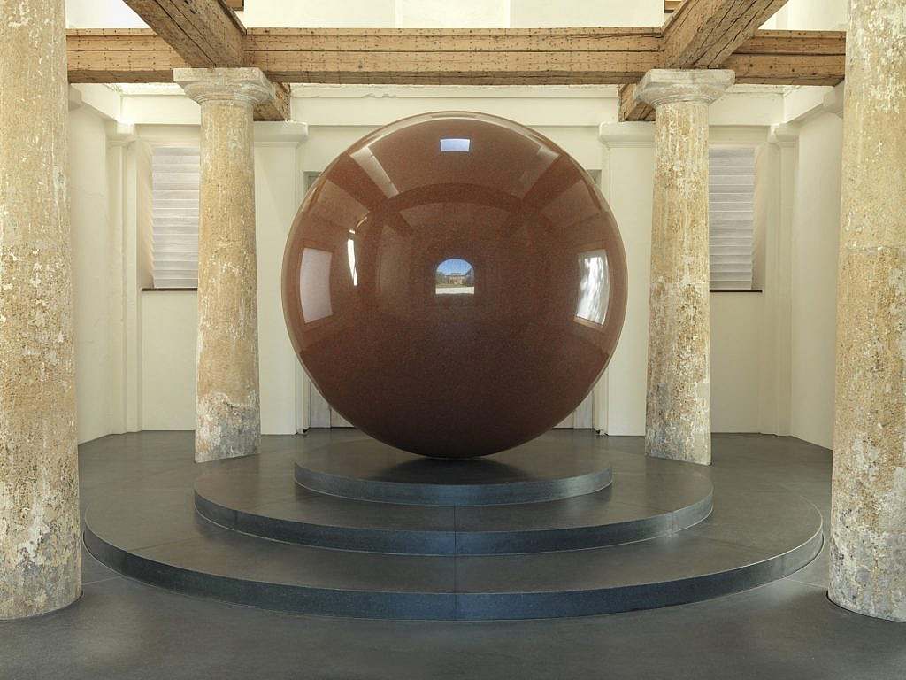 A large red sphere with a small arch in the centre, set on a three-tiered plinth and surrounded by four posts