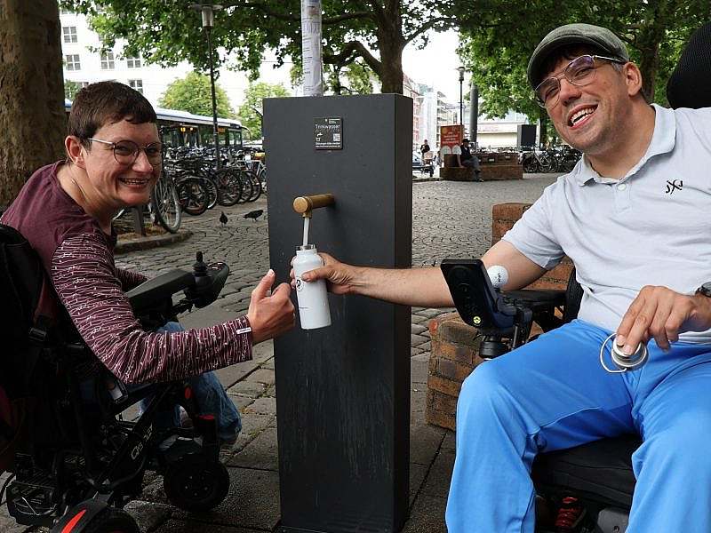 Zwei Personen im Rollstuhl holen sich Wasser am Brunnen